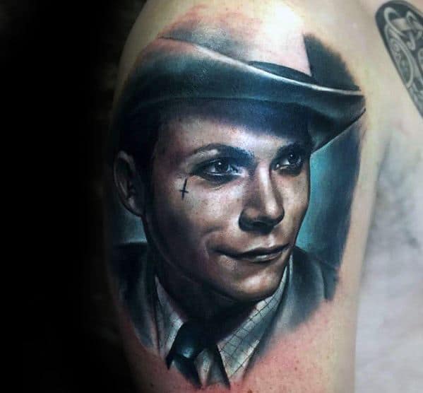 Tattoo of a man in a suit and hat with a small cross under his eye, on a person's upper arm