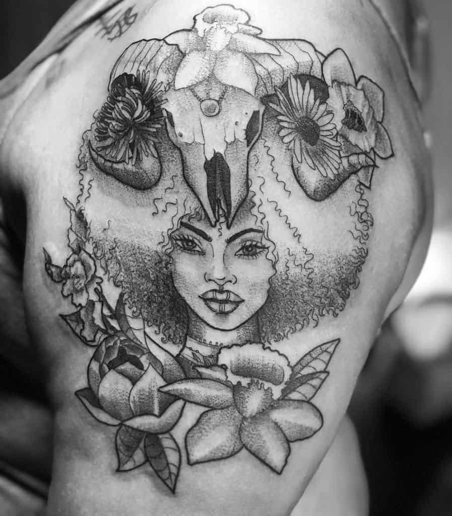 Black and white tattoo of a woman's face with a ram skull and flowers on the upper arm