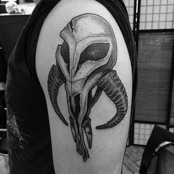 Stylized Mandalorian skull tattoo with a helmet-like top and detailed curved horns, inked in black and gray on the upper arm.