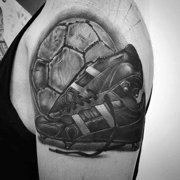 Realistic black and grey tattoo of a worn soccer ball and cleat on the upper arm, showcasing deep shading and texture details.