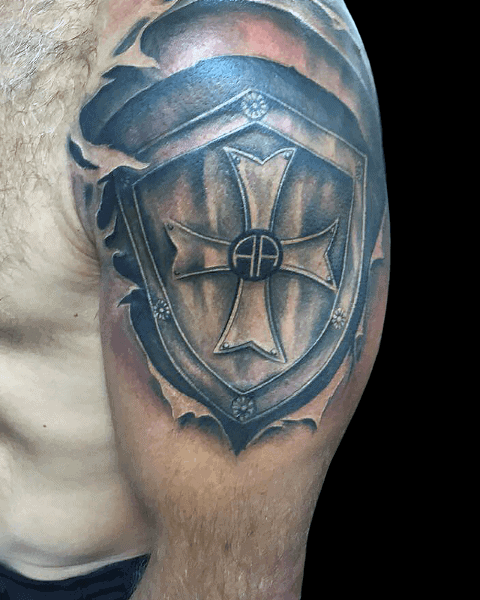 Upper arm tattoo of a 3D medieval-style shield with a bold cross and initials “AP” at the center, set against a torn skin effect.