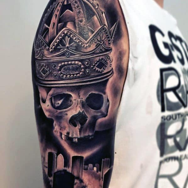 Tattoo of a crowned skull with a graveyard background on a person's upper arm