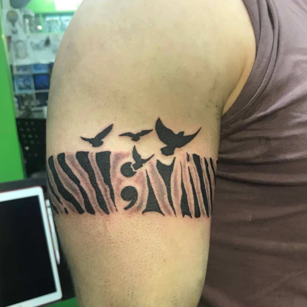 The tattoo features a semicolon surrounded by abstract shapes and flying birds, creating a powerful and dynamic design on the upper arm.