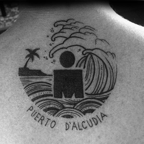 Ironman logo tattoo surrounded by waves, a palm tree, and the text "Puerto d'Alcudia" at the bottom, symbolizing a triathlon event at that location.