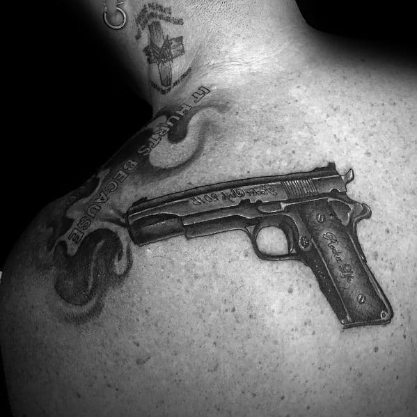 Realistic black and gray tattoo of a 1911 pistol on the upper back, featuring engraved details, smoke effects, and bold shading for depth.