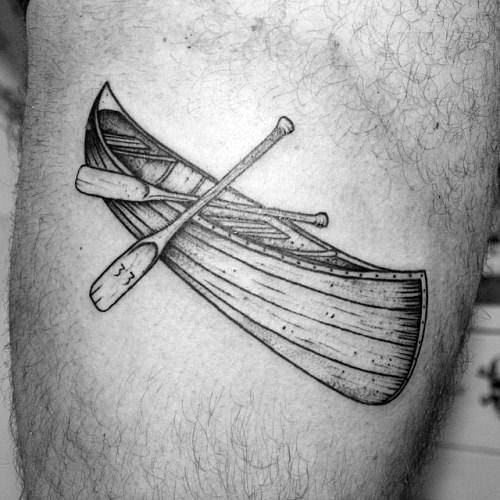 Alt text: Black and grey tattoo of a canoe with two crossed paddles, emphasizing a detailed and classic design.