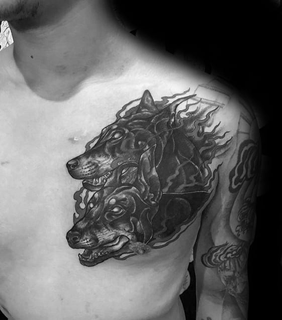Black and grey chest tattoo featuring two fierce Doberman heads with glowing eyes and flame-like accents, creating a bold and intense design.
