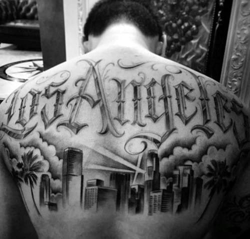 Upper back tattoo of Los Angeles skyline with palm trees, spotlights, clouds, and bold “Los Angeles” script across the shoulders.