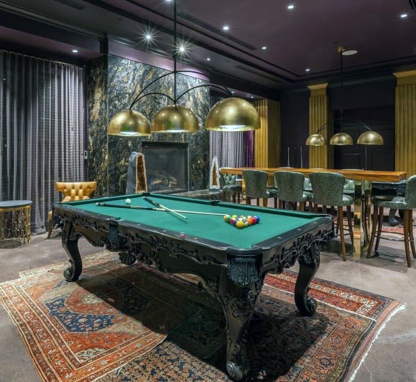 Upscale billiards room with ornate pool table, gold pendant lights, and marble fireplace backdrop.