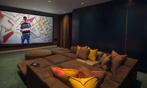 Home theater with a large screen showing a person spray painting, surrounded by brown couches and colorful pillows