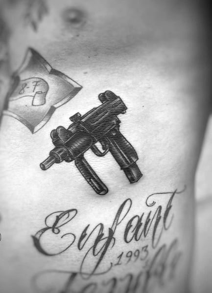 Realistic black and grey Uzi tattoo on the ribcage with fine shading and intricate details, placed above elegant script reading "Enfant Terrible 1993."