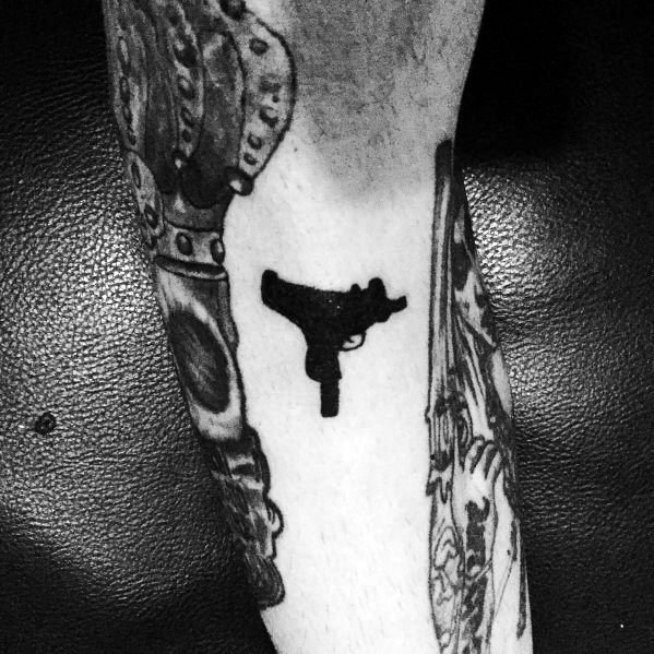Small black Uzi silhouette tattoo on the forearm with a minimalist design, contrasting against surrounding detailed tattoos.