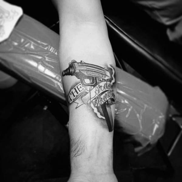Realistic black and grey Uzi tattoo on the inner arm with a banner wrapped around it reading "The Lord is Strength," blending firearm imagery with script.