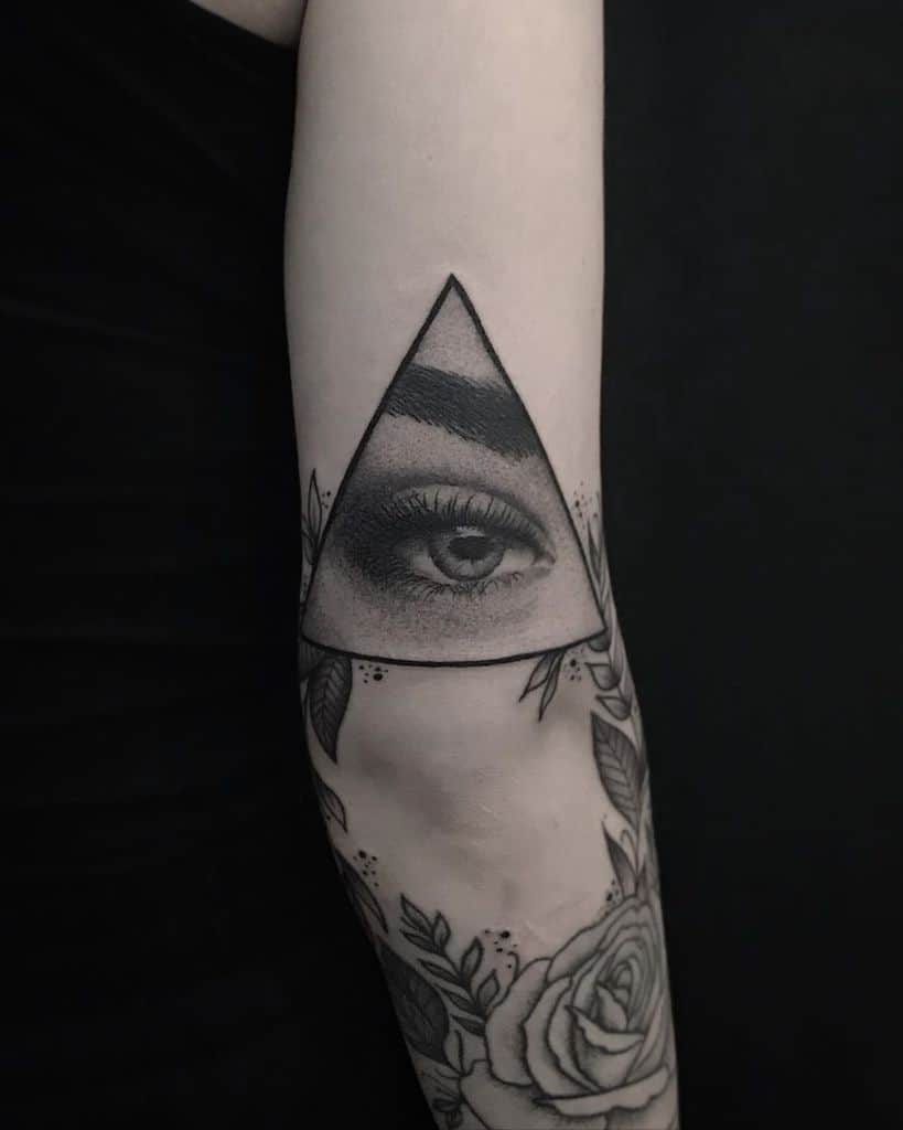 Tattoo of a realistic eye inside a geometric triangle, surrounded by floral elements, inked in black and grey on the forearm.