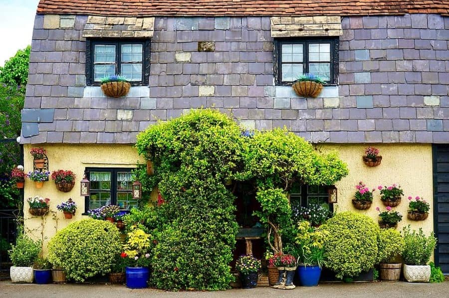 Cottage with a slate roof and colorful flowers adorning the walls, windows, and garden area, lush greenery surrounds the entrance