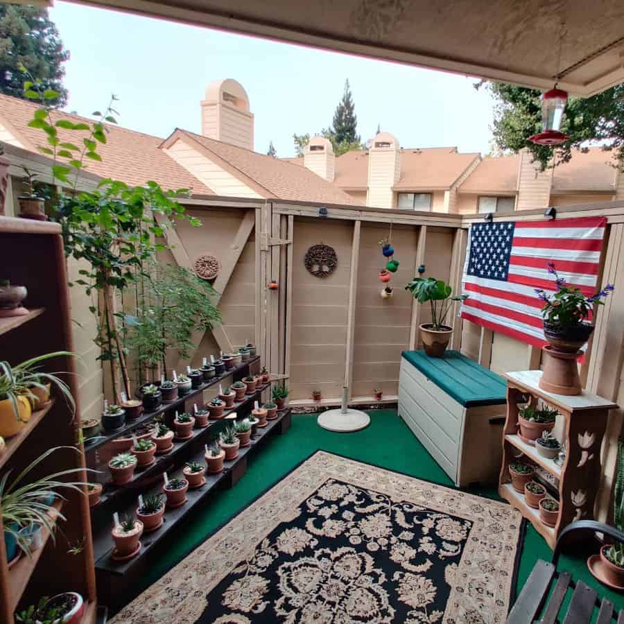 A cozy balcony garden with potted plants, a rug, a storage bench, and a U.S. flag on the wooden fence