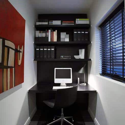 Compact home office with a computer, shelves, and books in a small room with a window and modern art on the wall