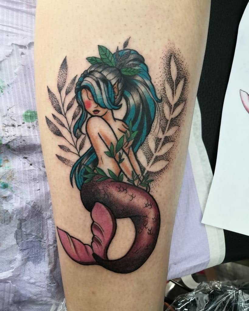 Colorful tattoo of a fantasy-style mermaid with blue hair, a pink and black tail, surrounded by leaves and intricate dot shading.