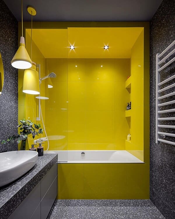 A modern bathroom with bright yellow tub walls, speckled grey surfaces, matching yellow pendant lights, and a white basin.