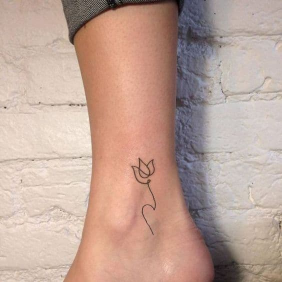 A minimalist black-line tulip tattoo on the ankle, featuring a simple flower with a curved, flowing stem.