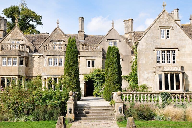 Grand Victorian-style mansion with ornate stone facade, gabled rooflines, tall chimneys, and lush landscaping featuring tall hedges
