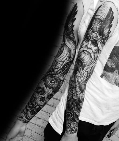 Black and grey full-sleeve tattoo featuring a Viking warrior with a winged helmet, detailed beard, skulls, and Norse mythology elements.