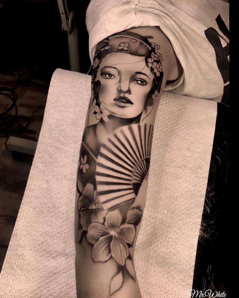 Black and gray forearm tattoo of a Geisha with delicate facial features, a detailed folding fan, and surrounding floral elements in a realistic style.