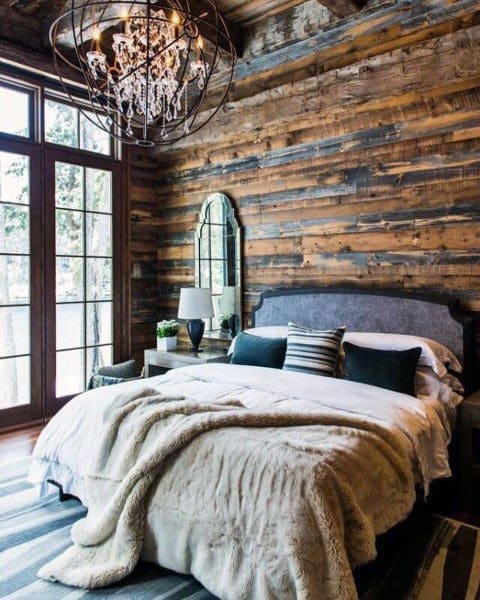 Rustic bedroom with wood-paneled walls, large bed, and chandelier by a window.