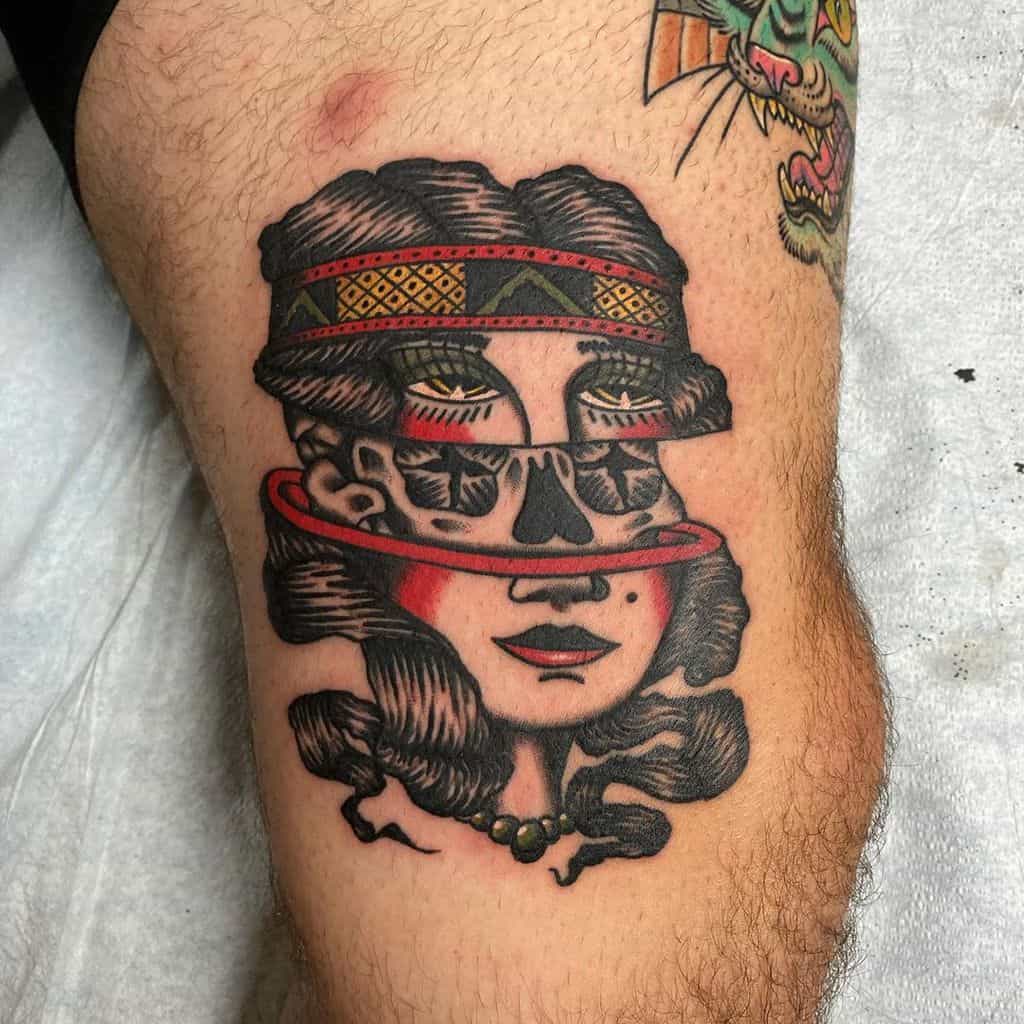 Traditional tattoo of a gypsy woman with a surreal split-face effect, revealing a skull beneath, featuring bold colors and intricate shading on the thigh.