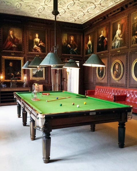 Traditional billiard room with green felt table, ornate portraits, and vintage lighting fixtures.