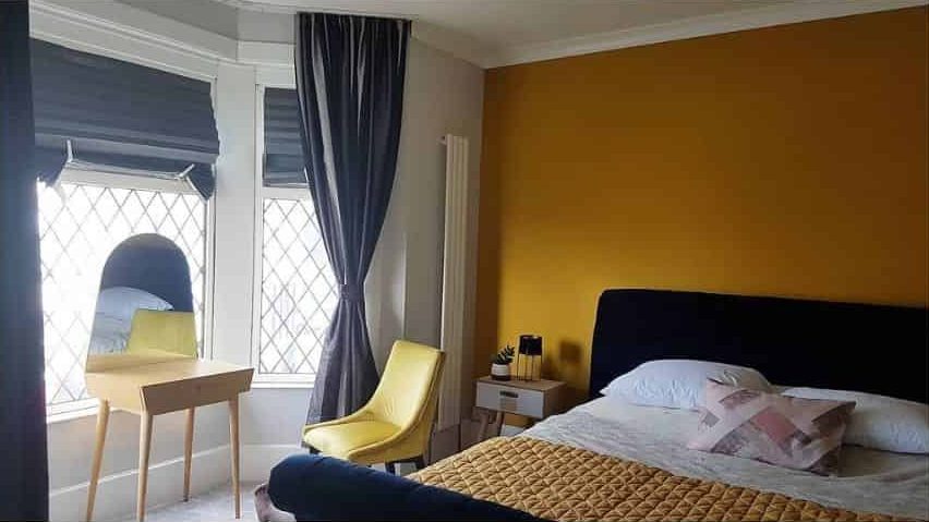 Modern bedroom with a yellow accent wall, plush bed, and bright yellow chair near leaded windows