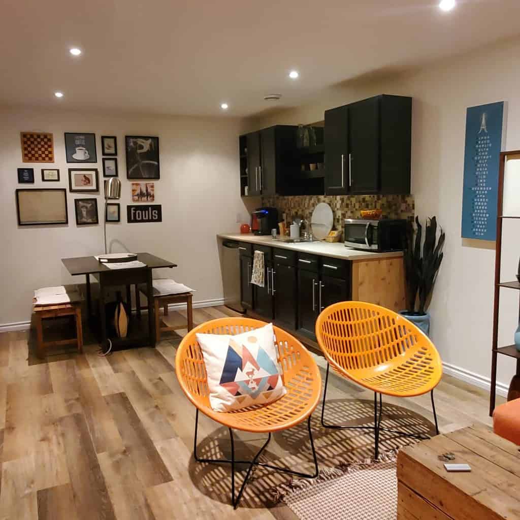 Modern basement with kitchenette, orange chairs, wall art, and wooden dining table on vinyl flooring.