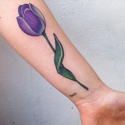 A bold purple tulip tattoo on the forearm, featuring rich shading, a long green stem, and an infinity symbol near the wrist.
