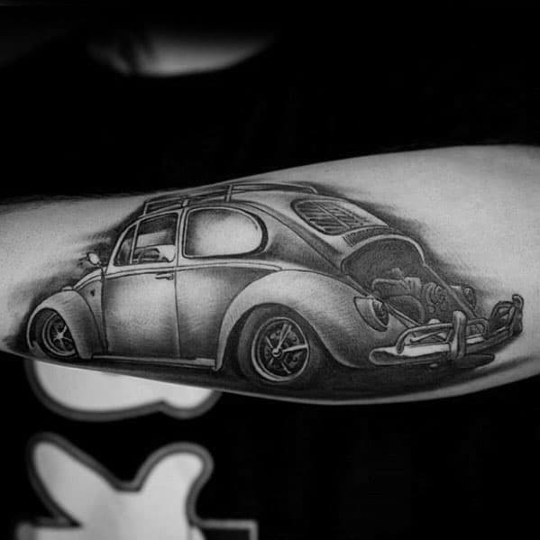 Detailed black and gray tattoo of a classic Volkswagen Beetle on the forearm, featuring a rear view with the engine exposed.