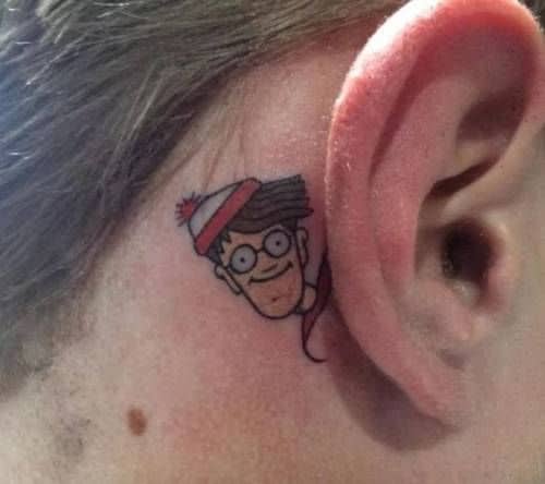 A small, colorful tattoo of a cartoon character wearing glasses and a striped hat, peeking out from behind the ear in a playful and subtle design.