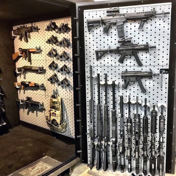 Walk-in gun safe vault with pegboard racks displaying rifles, shotguns, and pistols on walls and door.