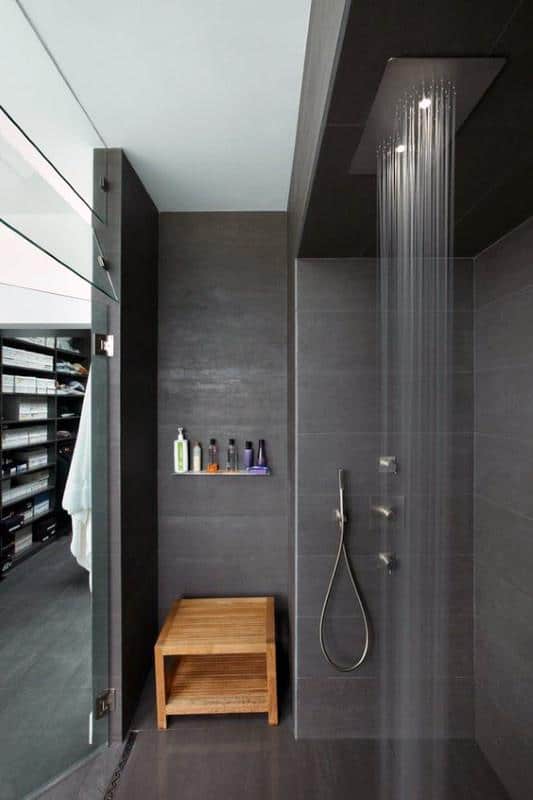 Modern shower with dark tiles, ceiling rainfall showerhead, wooden bench, and built-in shelf.
