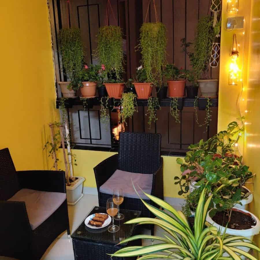 Small balcony with black wicker furniture, hanging plants, potted greenery, and string lights.