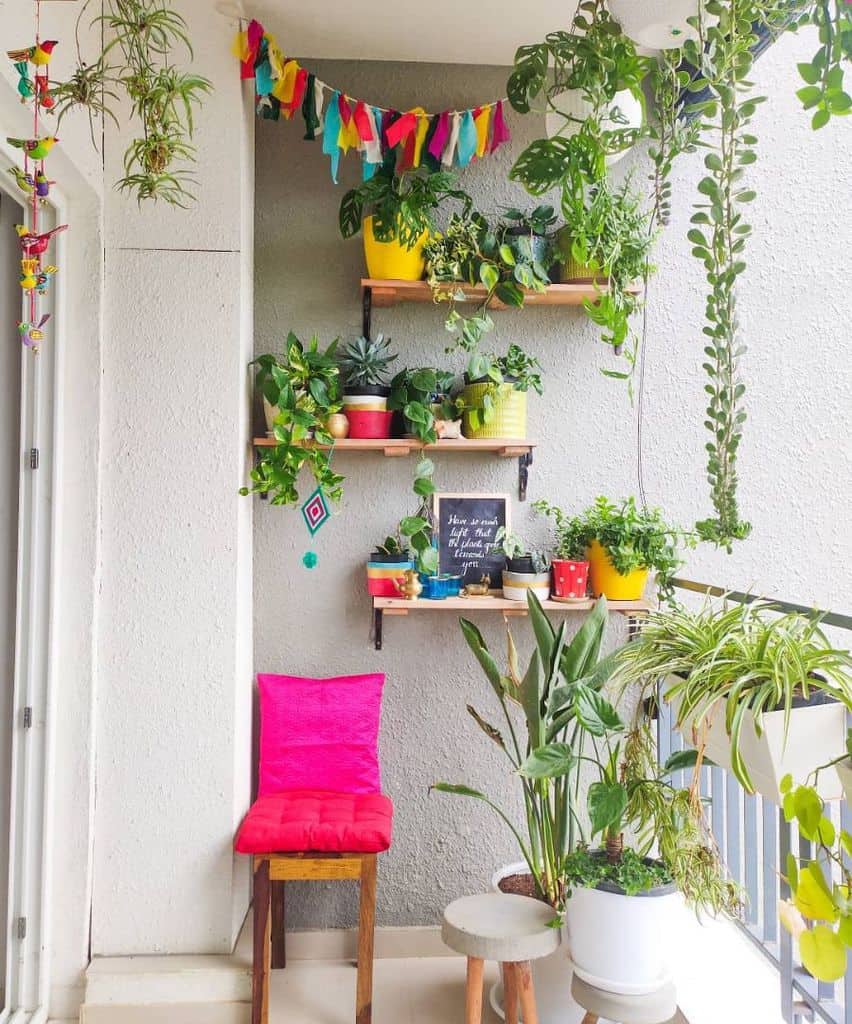 Colorful balcony with potted plants, hanging greenery, wooden shelves, and a pink cushioned chair.