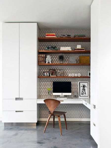 Minimalist home office with a wood chair, computer on a white desk, and shelves with books and decor against a patterned wall