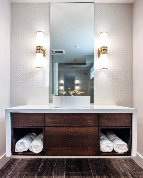 Modern bathroom vanity with gold wall sconces, large mirror, and rolled towels in shelves.