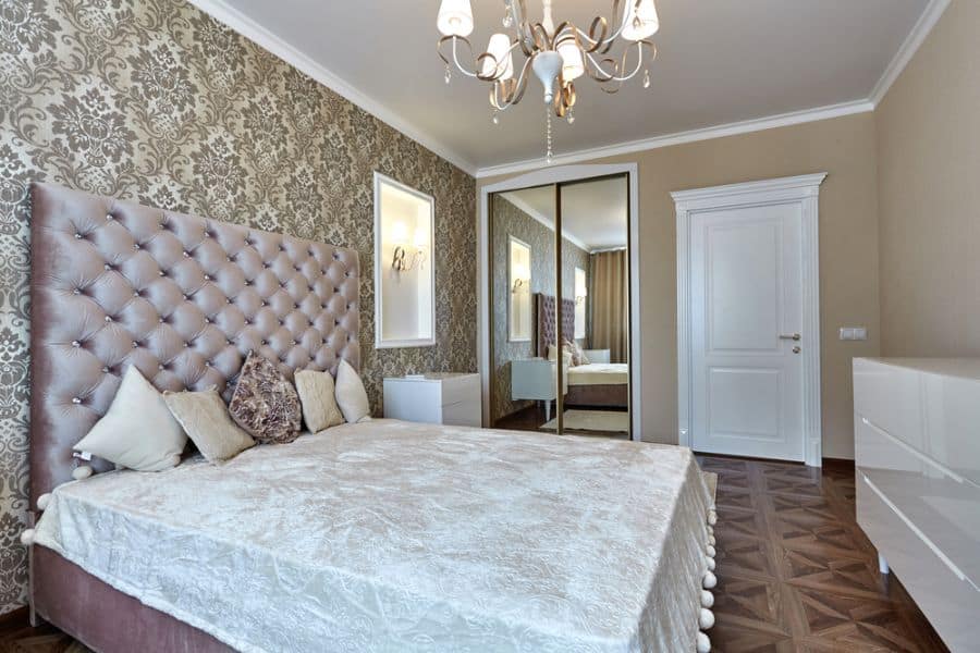 Elegant bedroom with a tufted bed, decorative wallpaper, mirrored closet doors, and a chandelier