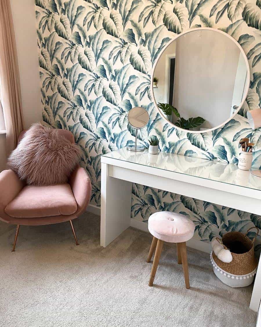 Stylish vanity area with leaf-patterned wallpaper, round mirror, pink chair, and small stool on cozy carpet
