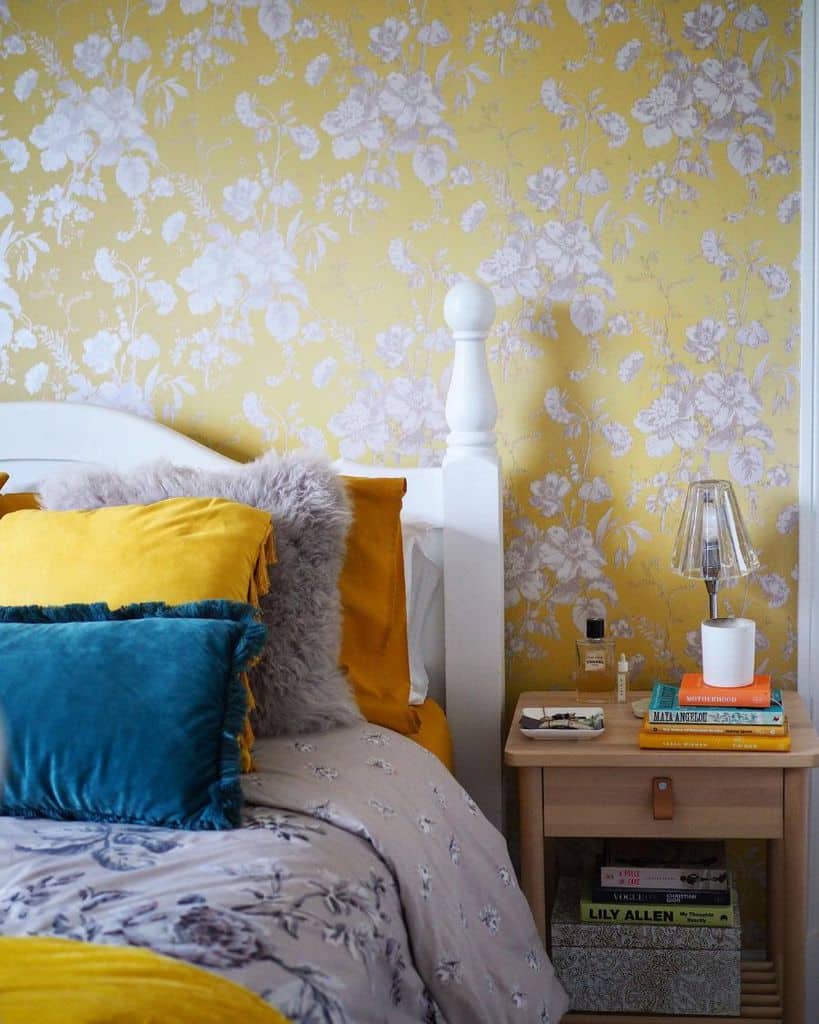 Sunny bedroom with yellow floral wallpaper, vibrant pillows on a white bed, and a nightstand with books and lamp