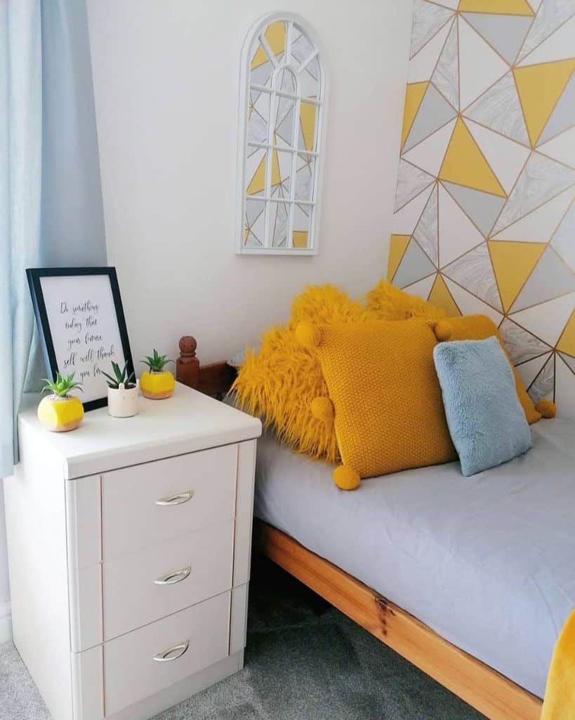 Cozy bedroom with vibrant yellow pillows, geometric wallpaper, and a white nightstand adorned with framed decor
