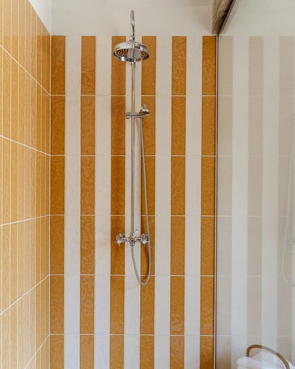 Bathroom featuring striped yellow-and-white tiles in the shower, a classic toilet, wicker basket, and glass partition.
