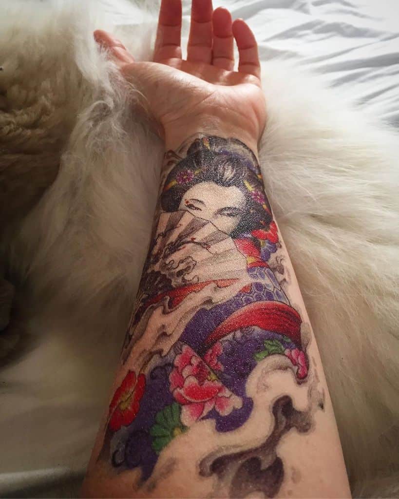 Vibrant forearm tattoo of a Geisha holding a fan, wearing a colorful kimono, surrounded by swirling clouds and floral elements.