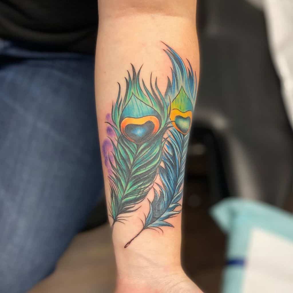 Colorful forearm tattoo featuring two peacock feathers with vibrant green, blue, and gold hues, detailed shading, and soft purple accents.
