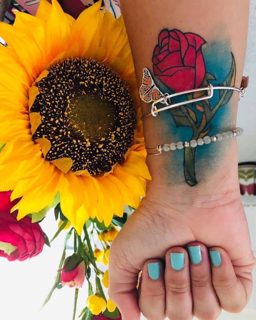 A colorful watercolor tattoo of a Beauty and the Beast rose, with bright red petals and soft blue accents