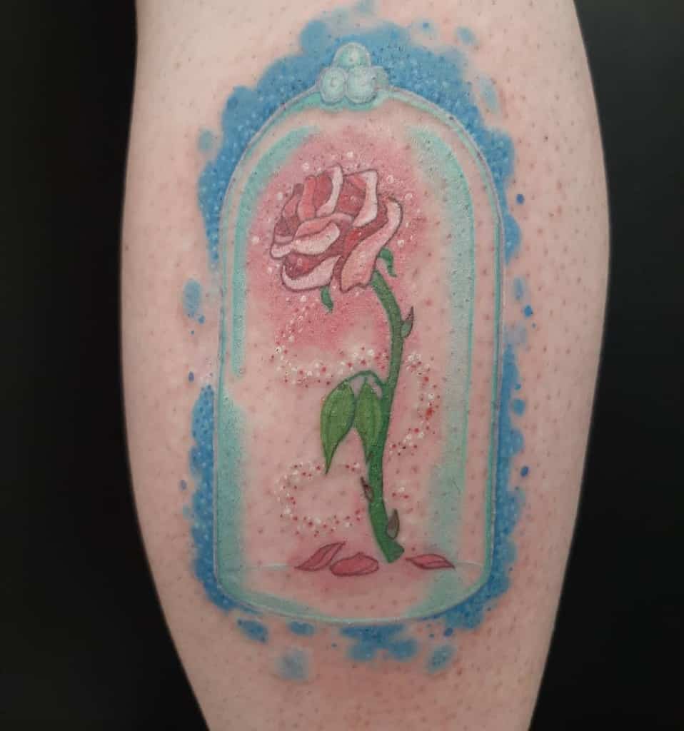 A watercolor tattoo of a pink rose inside a glass dome, with soft blue and pink accents and petals scattered below
